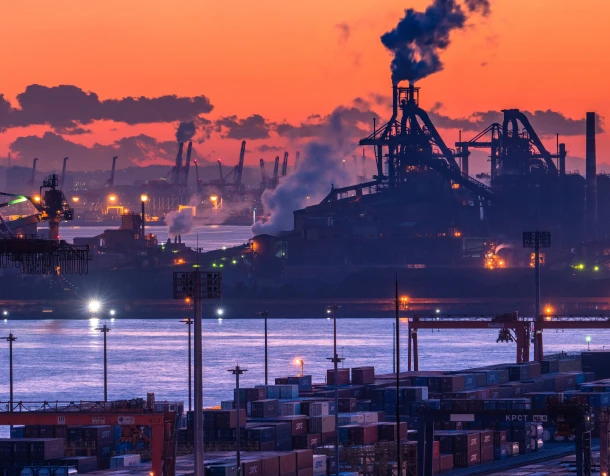 夕暮れの工場の写真。多くのプラントが集まり、煙突からは煙が吹き出し稼働している。